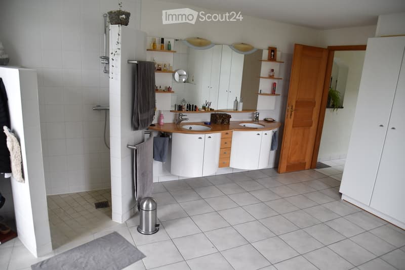White tiled floor, double sinks with wooden counter top, mirror, shelves, shower, towels, wooden door