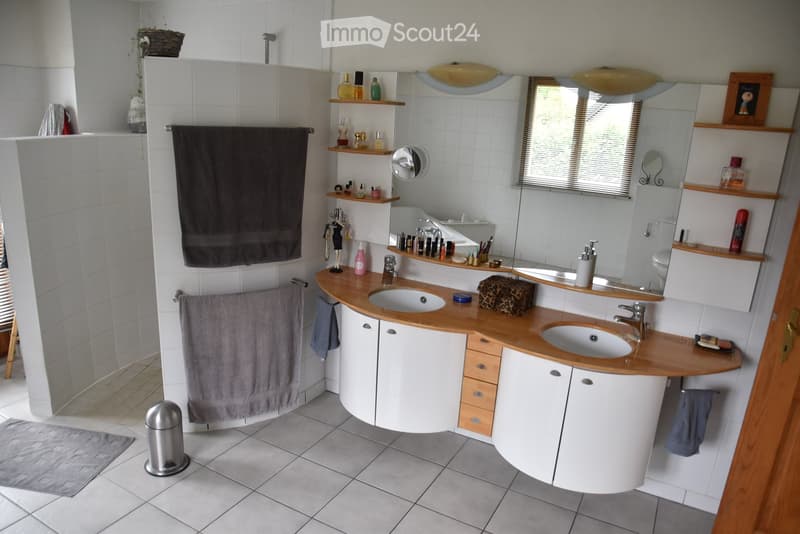 Bathroom: white tiles, wooden countertop, two sinks, mirror, wooden shelves, grey towels, waste bin, picture frame, walk-in shower, toilet