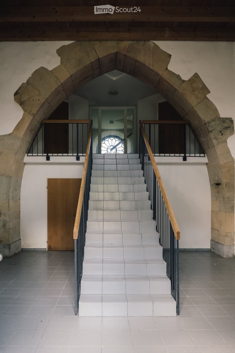 Stone building, arched entrance, white tiled floor, staircase with wooden handrails, metal railings on the upper floor