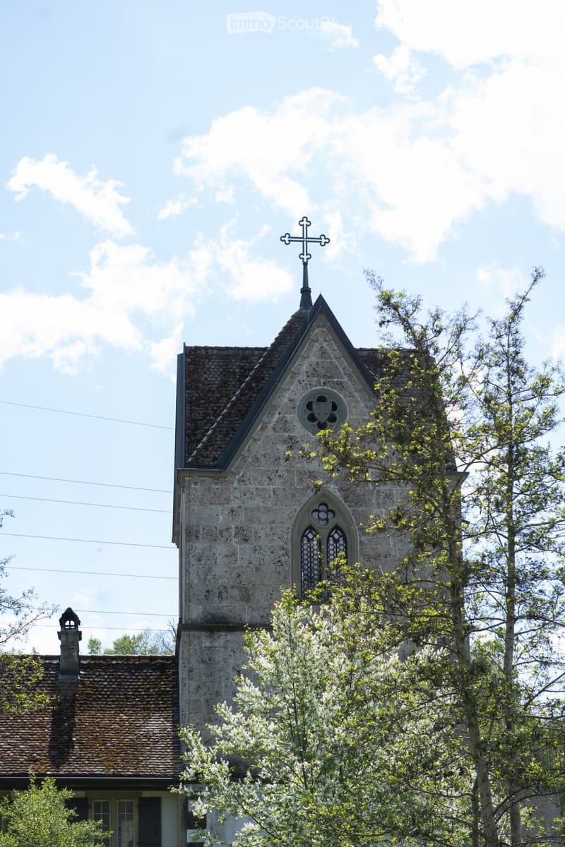Old stone building, cross on top, window with arch, trees in front
