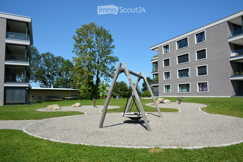 An outdoor space with gravel paths, swings, and grass. There are two residential buildings in the background.