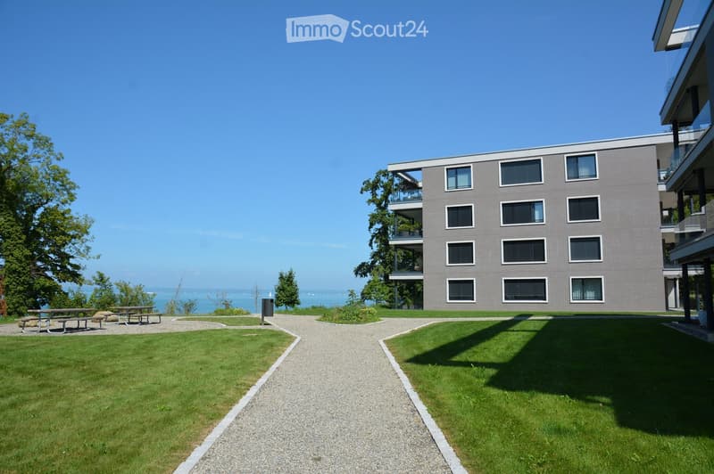 gray building, multiple windows, two benches, gravel path, grassy areas, view of the ocean