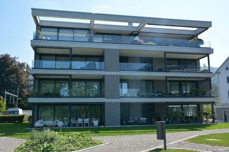 Modern multi-story building with balconies, elevated ground floor, glass windows, garden