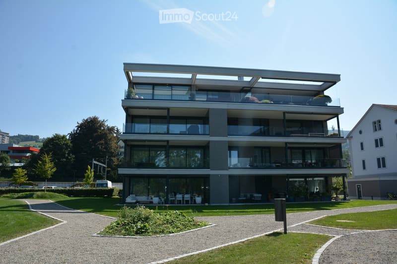 Modern apartment building, grey exterior, several balconies, raised ground floor, located in scenic area with green lawns and trees