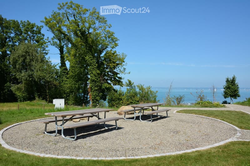Outdoor area with two picnic tables, seating, gravel pathway, surrounded by grass, distant ocean view