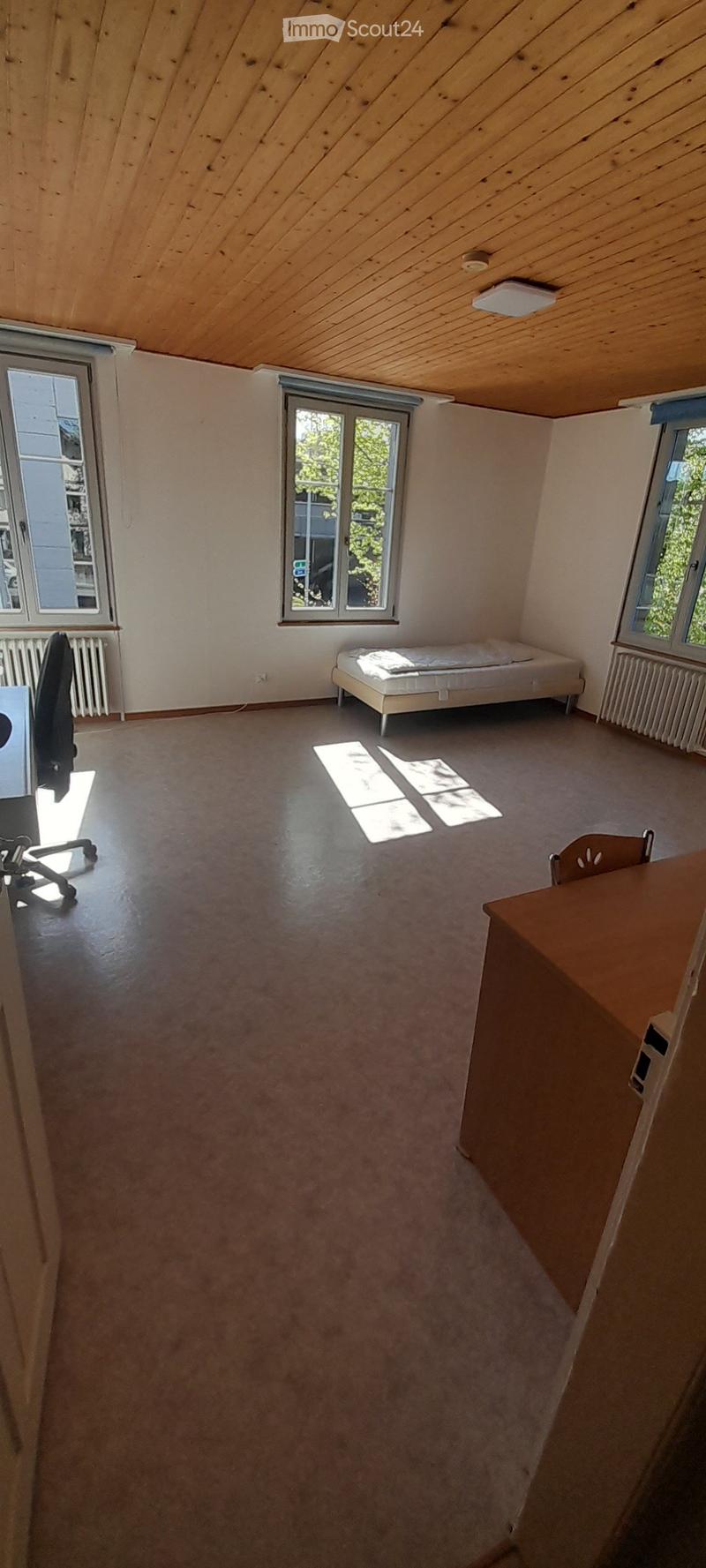 The image shows a room with a wooden ceiling, large windows, a bed, and a desk. The room appears to be a bedroom or a home office space.