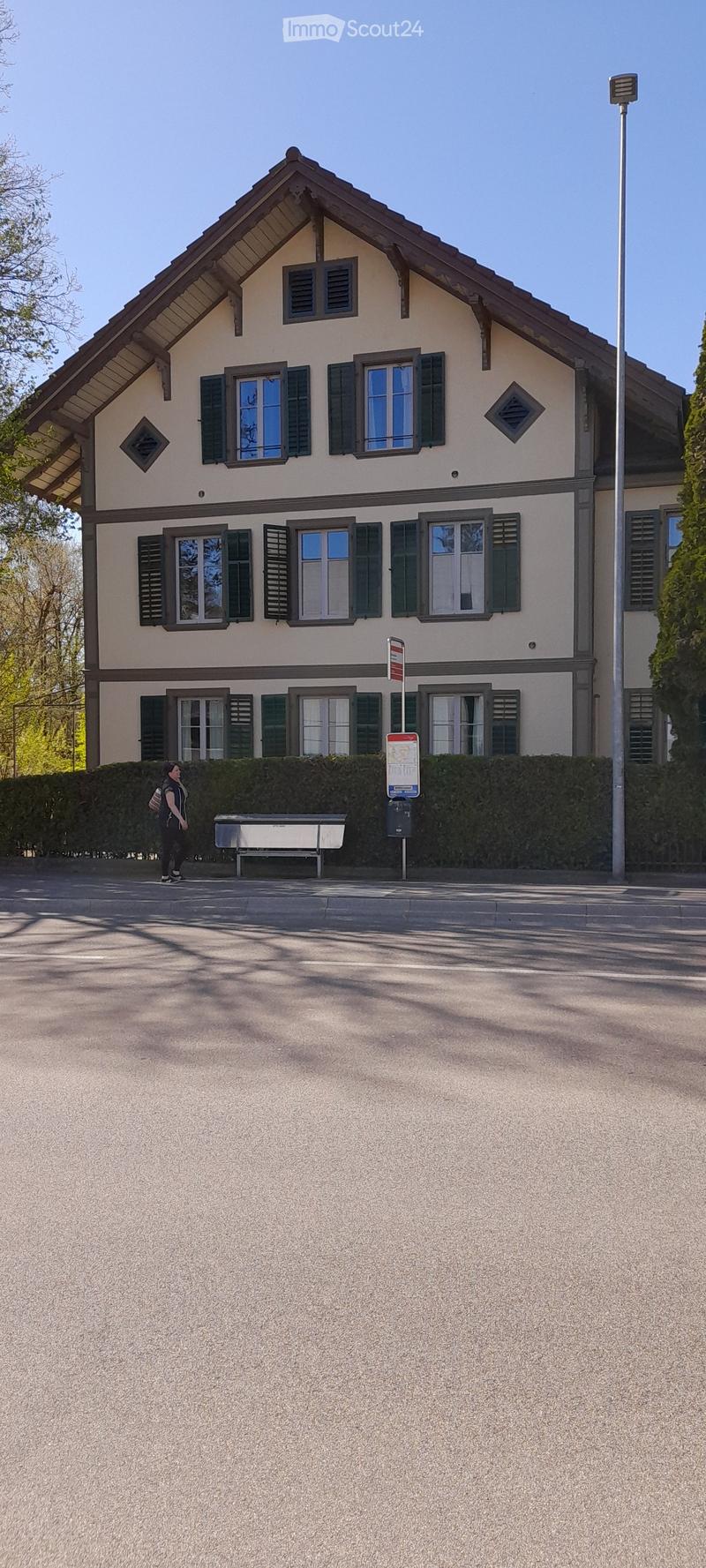3-story building with a traditional Swiss-style architecture, featuring a sloped roof, wooden shutters, and a balcony on the top floor. The building has a light-colored exterior and is surrounded by trees.