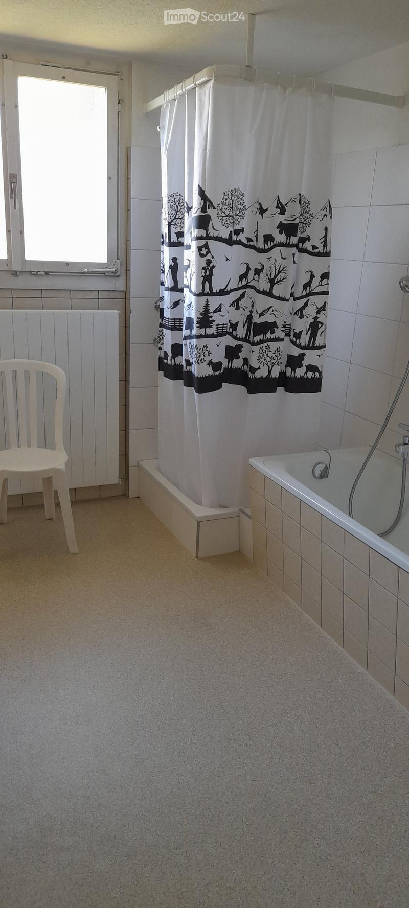 Tiled bathroom with a bathtub, shower curtain with a nature-themed design, and a white plastic chair.