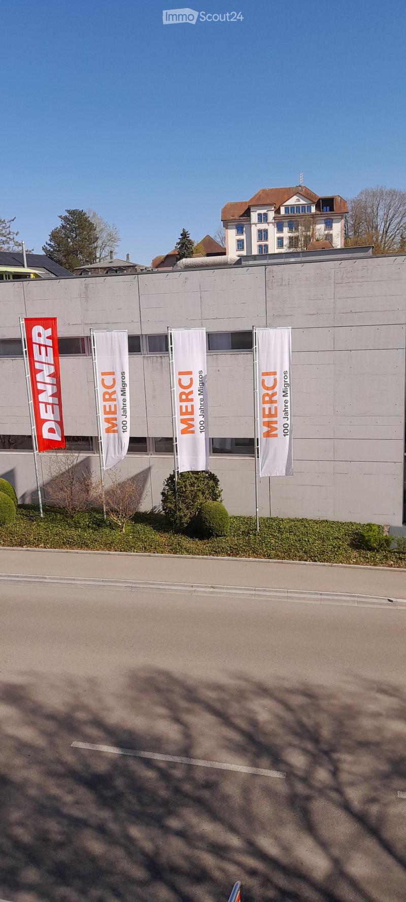 A multi-story commercial building with Denner and Merci banners displayed on the facade. The building appears to have a parking area in front of it.
