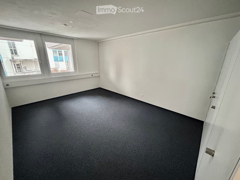 Empty room with white walls and ceiling, black floor, power outlet, windows overlooking buildings outside.