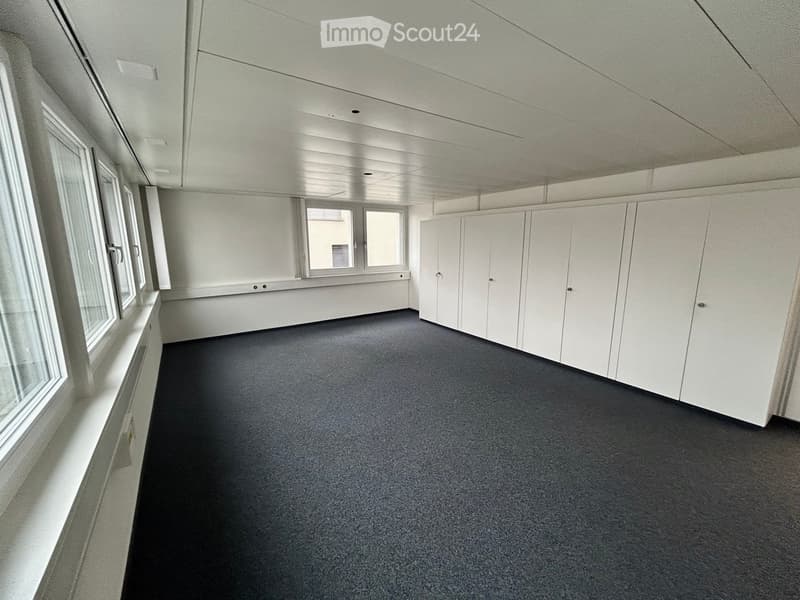 An empty office space featuring gray carpet flooring, white walls, multiple large windows, and built-in white cabinets against the wall.