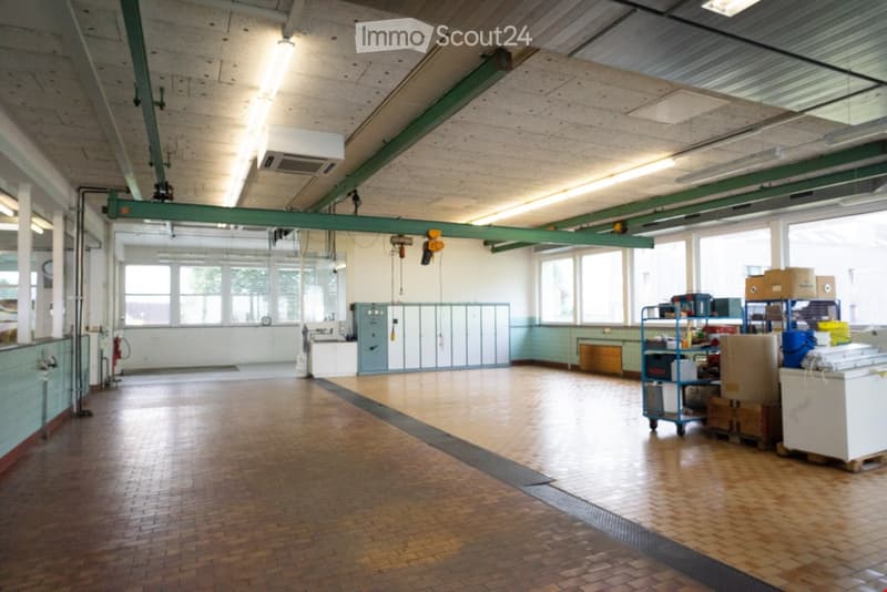 An empty room with tiled floor, steel beams and green columns on ceiling, two rows of windows, and some storage boxes and shelves on the right side.