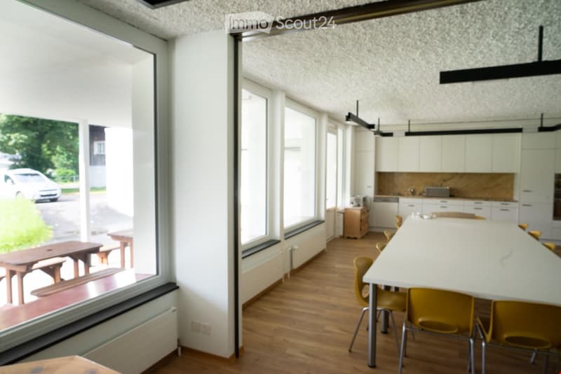 Dining area with large windows, white desk, yellow chairs, wooden benches, open kitchen area