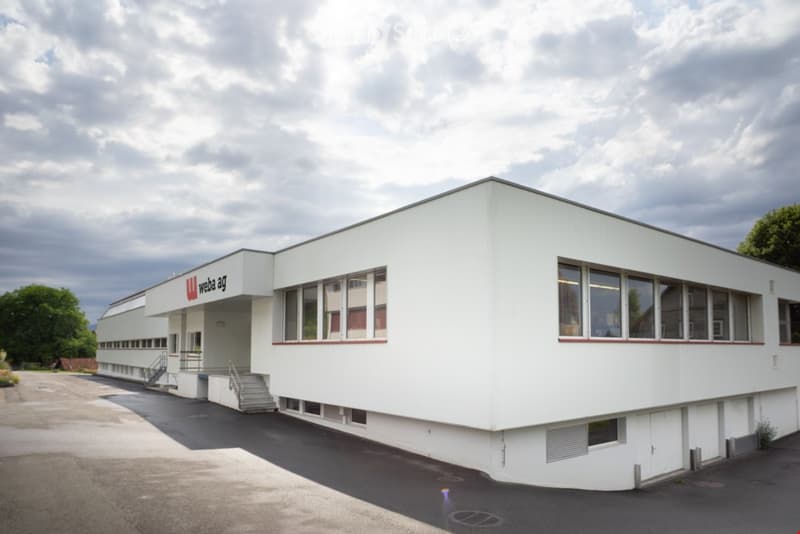 A large white building with glass windows, red accents, and a name sign 'weba ag' on the side. It has stairs leading to the entrance and is surrounded by a paved parking area with a tree in the distance. The building has a modern and sleek design with its