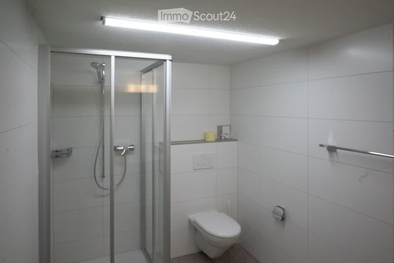 A small bathroom with white tiles and a glass shower stall, a white toilet, and a towel rack.