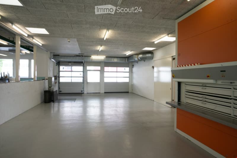 An empty building with white walls, grey floor, glass windows, two big doors on the back, and orange equipment on the right side.