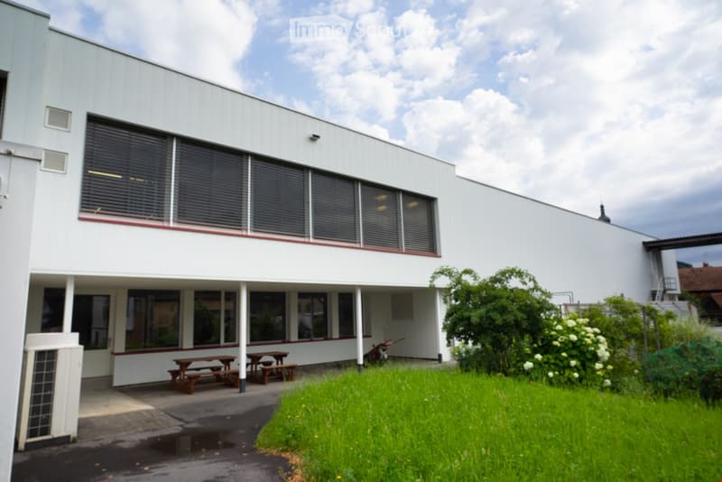 The building has an elevated ground floor. It has a modern, minimalist design with large windows and dark gray shutters. There are two wooden picnic tables in front of the building.