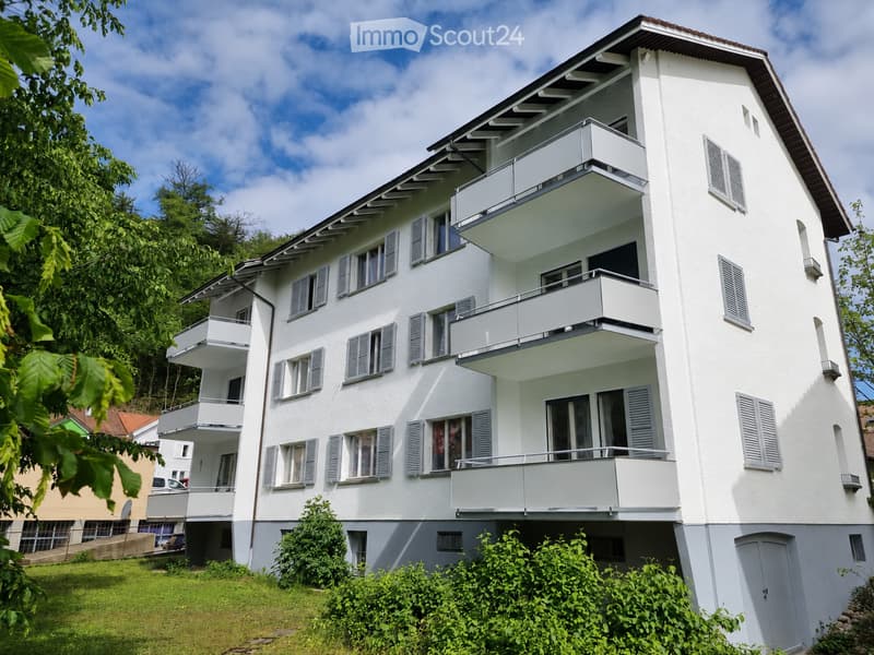 Building. A modern, white multi-story residential building with multiple balconies and large windows, surrounded by greenery.