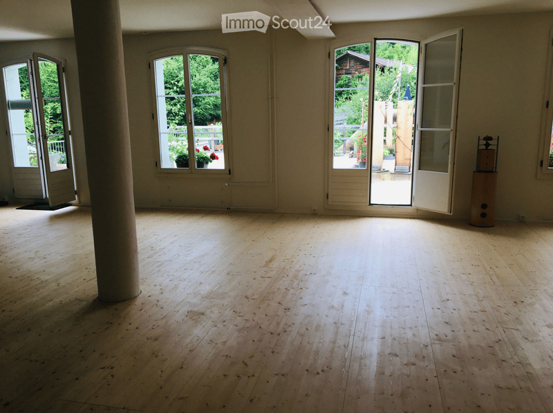 An empty room with wooden floors, white walls, and large windows. There are three doors, one of which is open, and a small wooden pillar in the center.