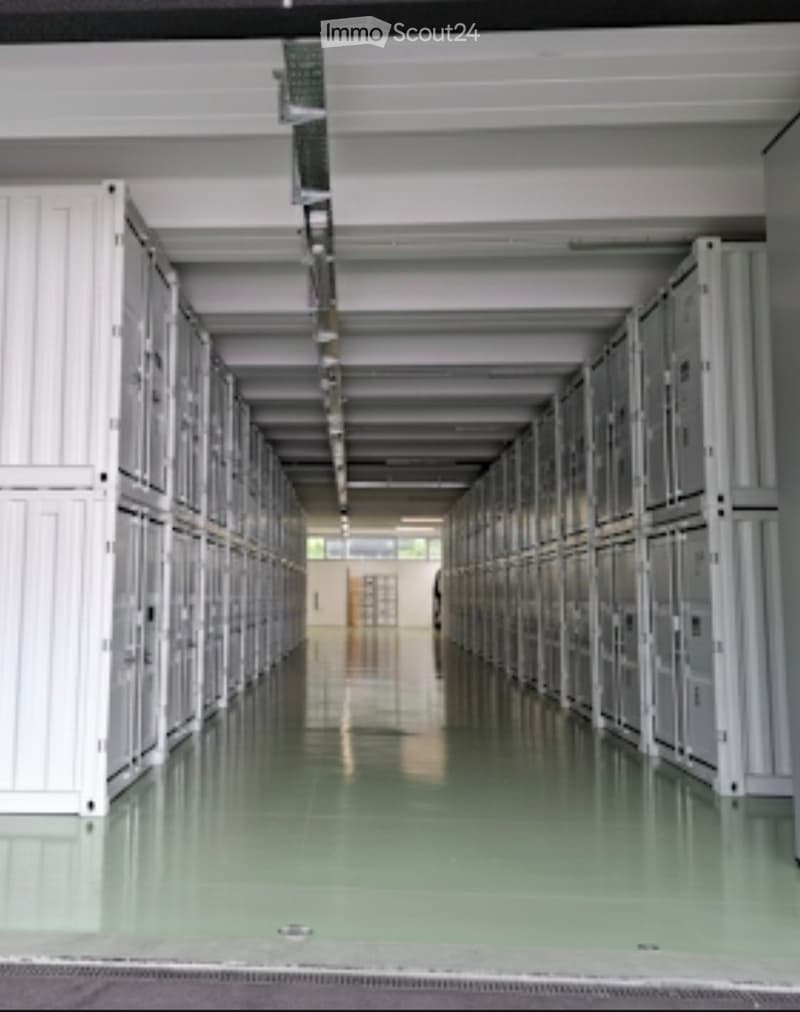 Storage facility with rows of storage units, white walls, green floor, metal beams, and windows at the end.