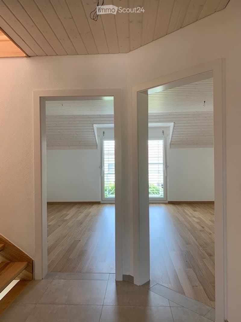 The image shows an empty room with white walls, a wooden ceiling, and hardwood floors. There are two windows visible, providing natural light to the space. The room appears to be a living room or bedroom, with a clean and minimalist design.