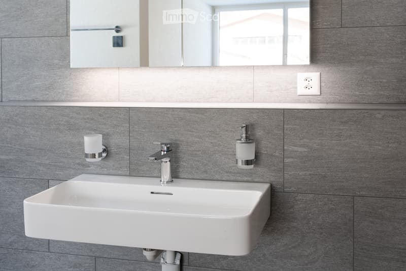white sink, faucet, mirror, soap dispenser, power outlet, gray tiles