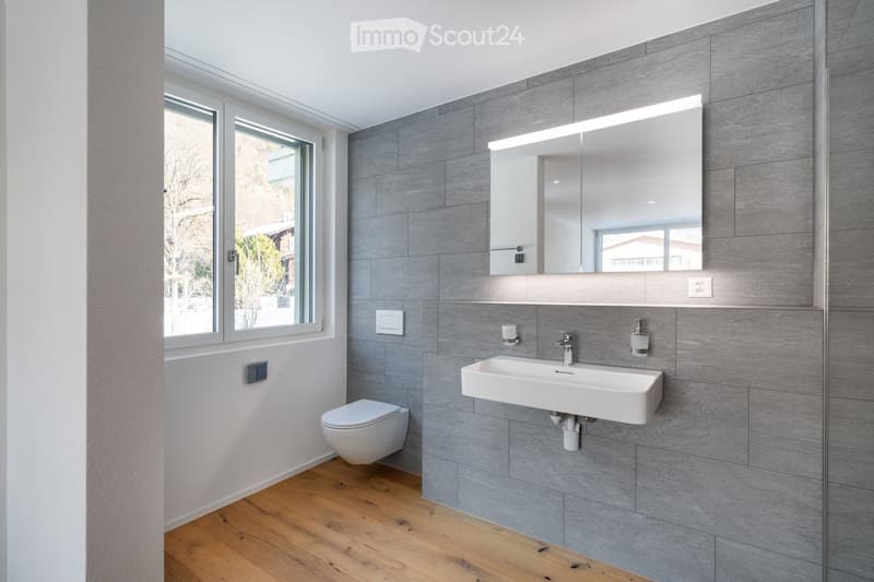 A modern, minimalist bathroom with a wall-mounted toilet, large mirror, silver fixtures, and wooden floor. Natural light comes through a window.