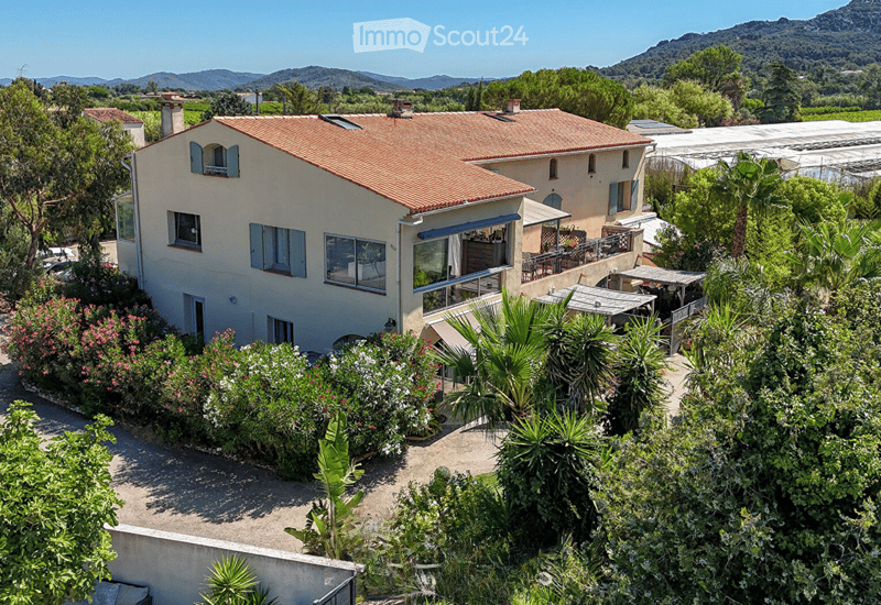 Two story house, red roof, balcony, surrounded by plants, palm trees, garden with flowers