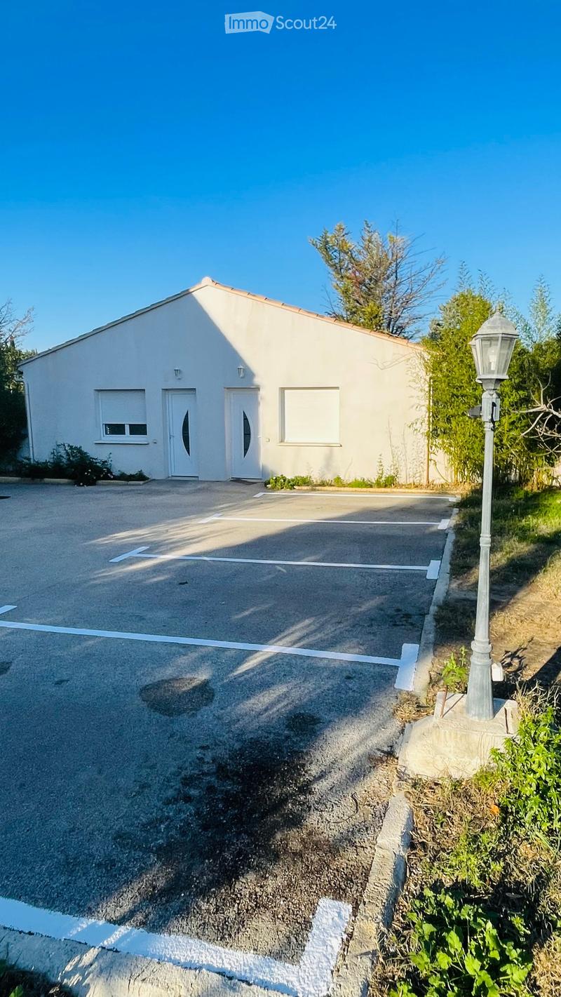 A white single-story house with a red tile roof. There are three doors on the facade, two large double doors and one smaller one on the left. There is a large parking lot in front of the house. There is a lamp post on the side of the parking lot. There ar
