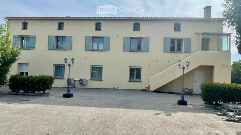 2-story beige house with multiple windows, concrete staircase, two street lamps, air conditioning units, spacious driveway, green shrubs