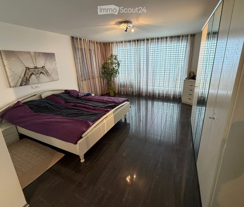 Bedroom with a bed, wooden floor, white walls, large windows, potted plant, and wardrobe.