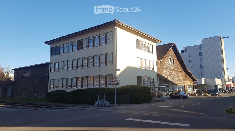 Two-story office building with glass windows, brown wooden exterior at the back, metal railing, trash bins, street light, cars parked, road, green grass, small bushes, and train tracks.