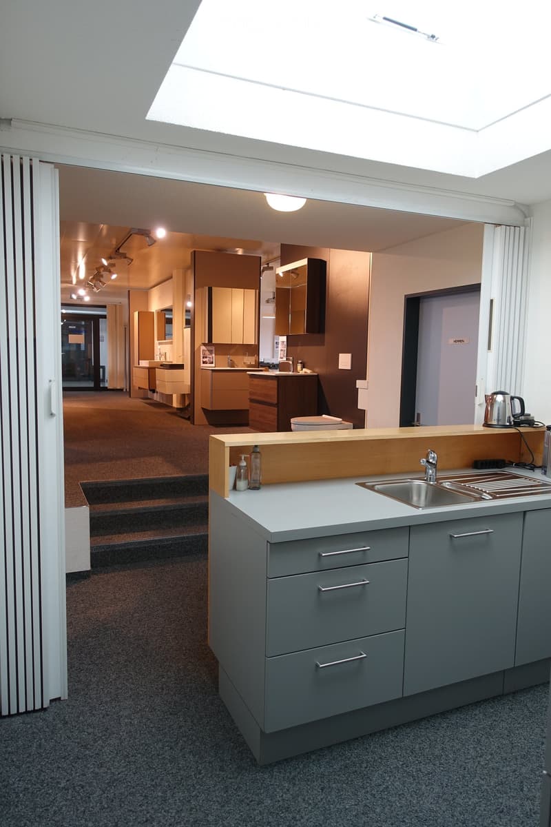 Open kitchen space with stainless steel sink and countertop, wooden cabinets, ceiling lamp, and tiled floor.