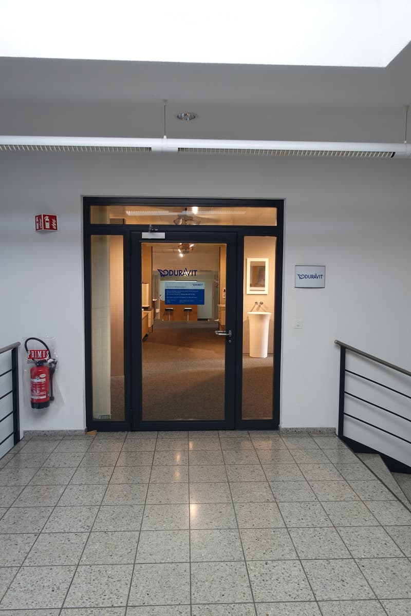 Main entrance to a modern office building with black doors, a glass entrance wall, white tiles on the floor, a red fire extinguisher, and a fire alarm on the wall.