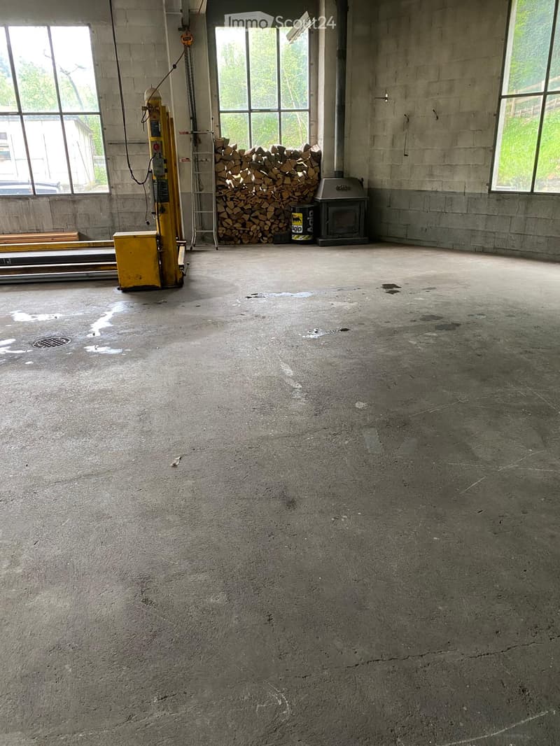 Empty room with concrete floor and walls, large windows, and a small fireplace with a stack of firewood nearby.