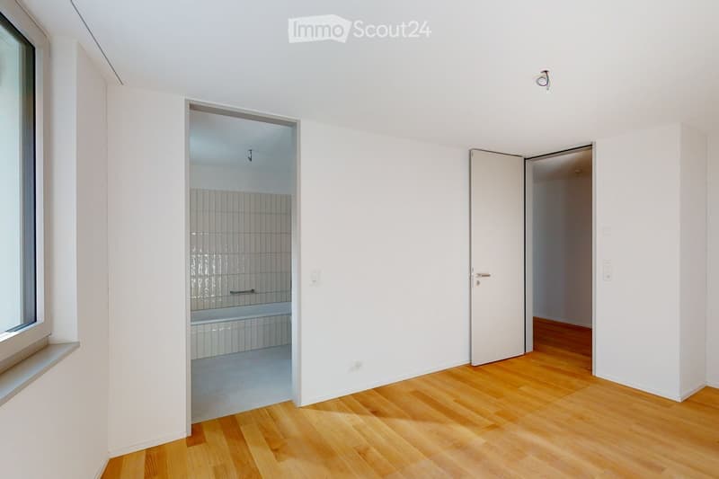 Empty room with white walls, wooden floor, a bathtub with glass door, and an open door leading to another room