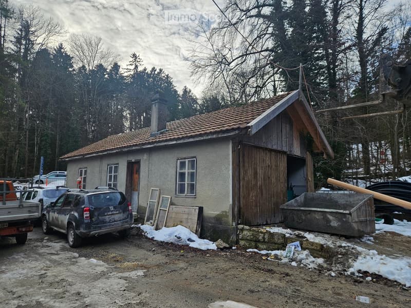 Small house made of wood and stone with a chimney, parked cars, snow-covered ground, wooden structure, trees in the background.