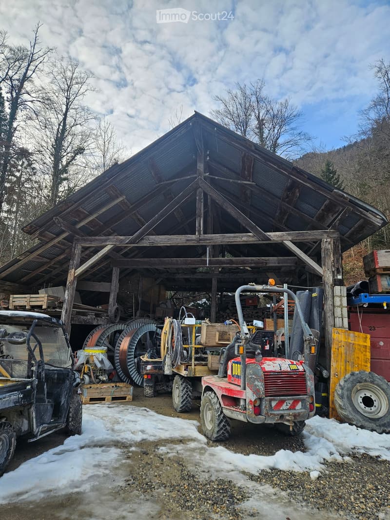 Barn with wooden trusses, two vehicles parked in front, equipment and tools inside, snow on the ground, metal storage units, large tires, storage boxes.