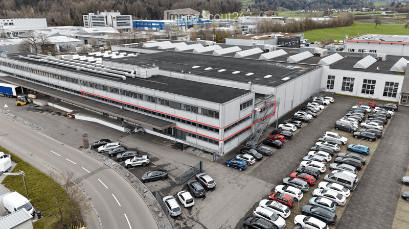 Aerial view of a large industrial building with a flat roof, surrounded by a parking lot filled with numerous cars. The building has multiple sections and is situated in a rural area with other structures and open fields in the background.