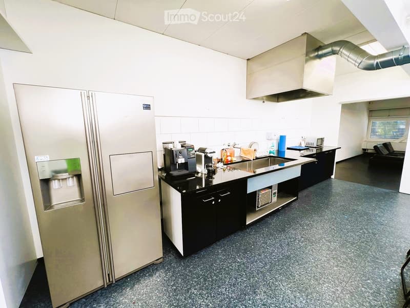 An empty kitchen with a refrigerator, black countertops, and cabinets, with a coffee machine, sink, and stove hood.
