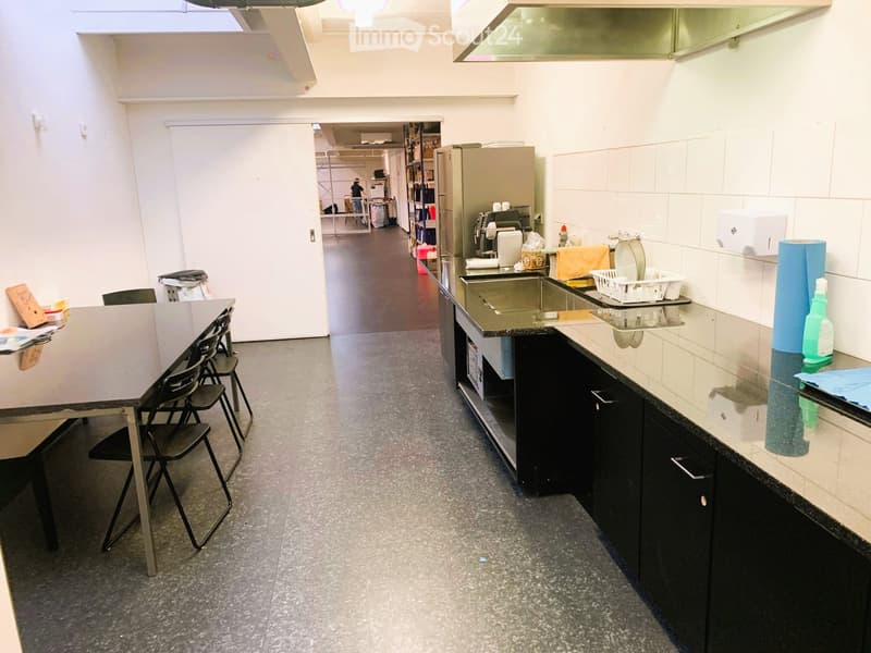 A rectangular kitchen room with black countertops, black cabinets, grey flooring, a stainless steel refrigerator, a stainless steel double sink, two folding tables with chairs, and a white door leading to another room.