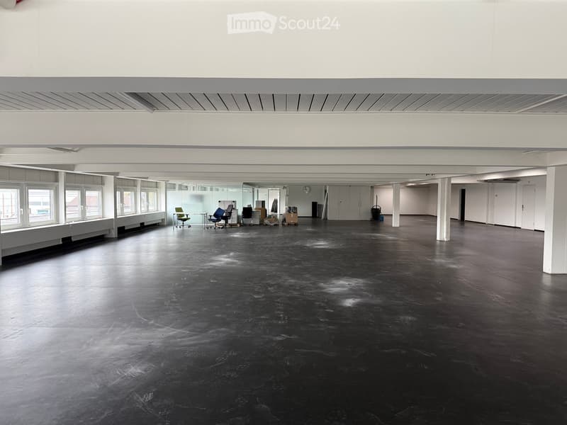 Open-plan room with dark polished concrete floor, large windows, white walls, and minimal furniture including chairs and a small table.
