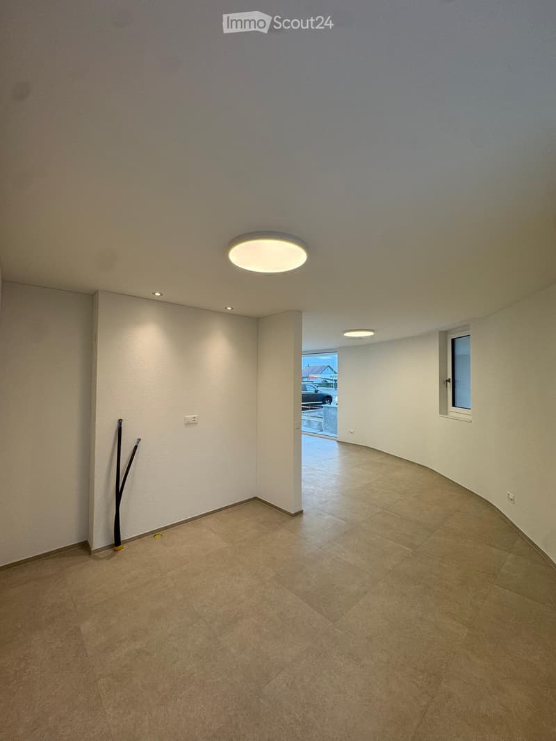 An empty room with white walls and tiled floors, has windows and ceiling lights, there is a car parked outside