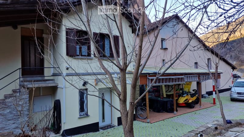 Raised ground floor, beige exterior walls, windows with brown shutters, covered porch, covered parking area, a car parked outside