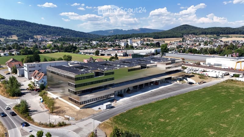 Aerial view of a modern industrial building with solar panels on the roof, surrounded by a parking lot, greenery, and a town in the background.