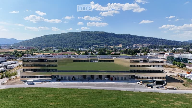 Modern multi-story building with large windows and solar panels on the roof, surrounded by a parking lot and greenery with a mountainous backdrop.