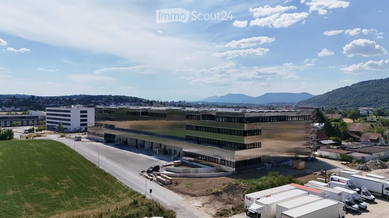 Aerial view of a modern industrial building with a large parking area, several trucks, and a surrounding landscape with hills in the background.