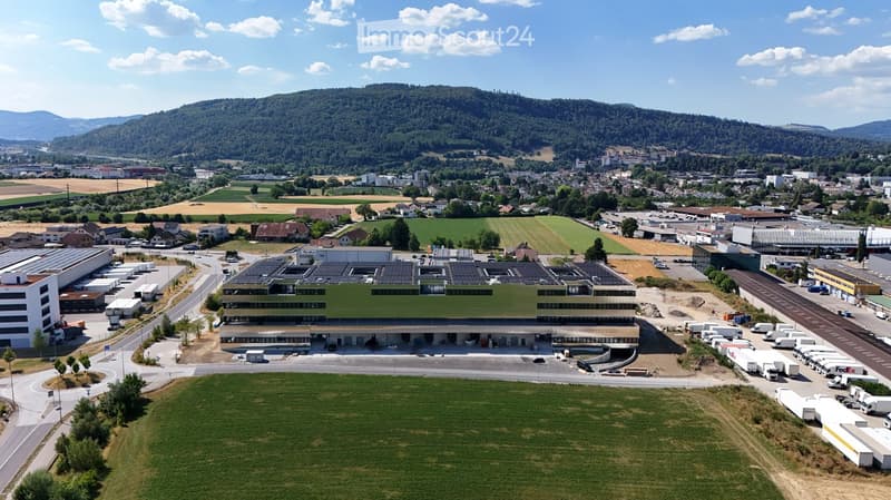Aerial view of a large industrial building with solar panels on the roof, surrounded by parking lots, trucks, and a rural landscape with fields and hills in the background.