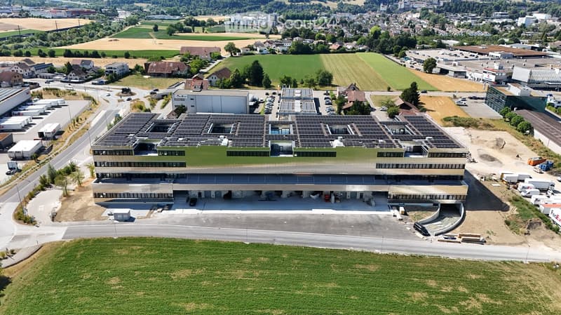 A modern, multi-story building with a green roof and solar panels, surrounded by a parking lot and industrial area.