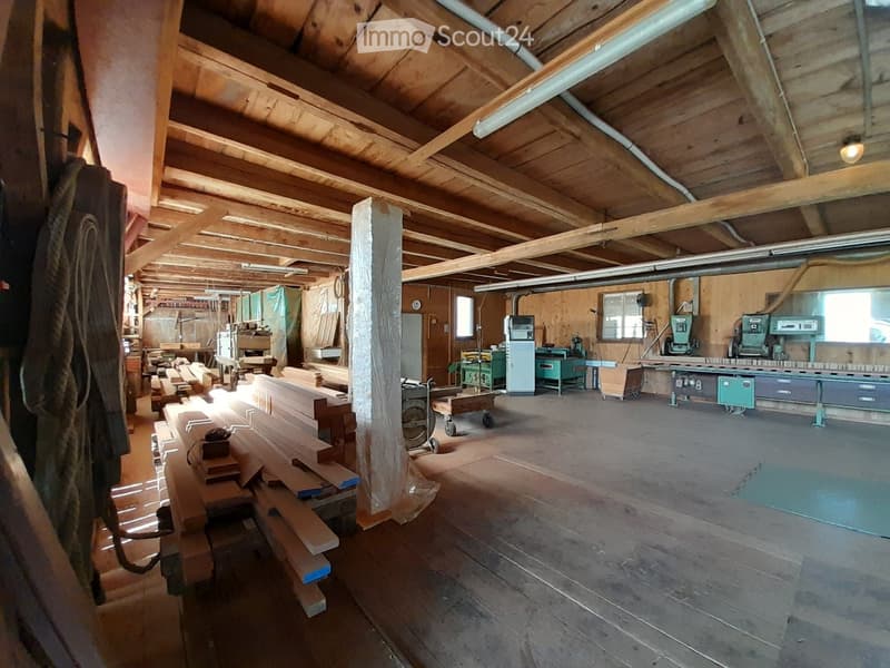 Workshop with wooden beams, concrete floor, various woodworking tools, machinery, and materials arranged, sunlight filtering in